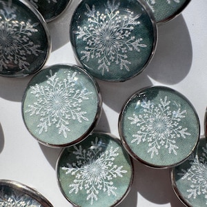 May include: A collection of circular buttons featuring a dark green fabric background with a white, intricate snowflake pattern printed on each. Each button is encased in a smooth, clear, rounded glass-like dome with a polished silver metal rim.