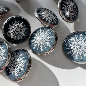 May include: A collection of round buttons featuring a dark blue fabric background with a white embroidered snowflake pattern. Each button is encased in a polished silver-toned metal rim and includes a small metal loop on the back for attachment.