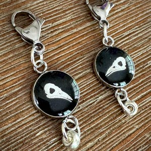 May include: Two silver-toned charm pendants with a black background and a white bird skull design. Each pendant is round and attached to a silver clasp. The pendants are displayed on a wooden surface.