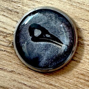 Puede incluir: Un colgante redondo plateado con un diseño de calavera de pájaro negro sobre un fondo gris moteado. La calavera es una silueta detallada, y el colgante tiene una superficie brillante y abovedada. El colgante está sobre una superficie de madera.