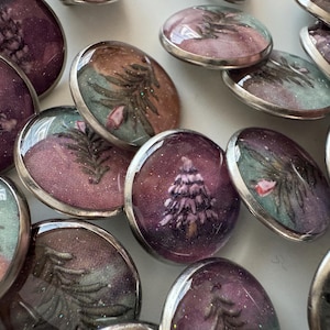 Peut inclure: Une collection de boutons ronds et bombés avec des bordures en métal argenté. Les motifs des boutons présentent des peintures miniatures de paysages dans des tons de violet, de vert et de marron, sur le thème de l'hiver ou de la forêt. Certains boutons représentent un arbre enneigé.
