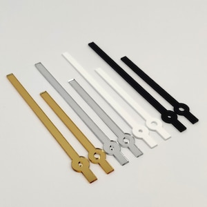 May include: Assortment of clock hands in gold, silver, and black. The hands are made of metal and have a circular shape at the end.