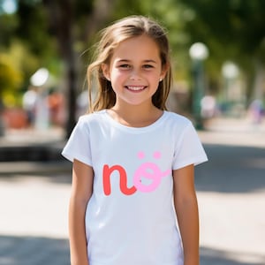 Pode incluir: Uma camiseta branca com gola redonda com a palavra "në" em vermelho e rosa. A camiseta é usada por uma pessoa jovem com cabelo longo e castanho claro, sorrindo ao ar livre. O fundo está desfocado com árvores verdes.