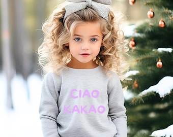 May include: A young girl with blonde curly hair wears a gray sweatshirt with the words "Ciao Kakao" printed in purple. She is wearing a silver glitter bow headband.