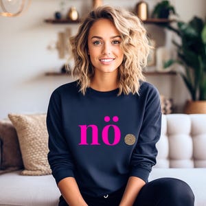 Puede incluir: Una sudadera azul marino con la palabra "nö" en rosa brillante, acentuada con un círculo con estampado de leopardo. La sudadera es de manga larga y tiene cuello redondo. La modelo sonríe.