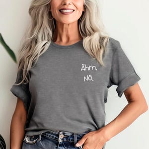 Può includere: Una maglietta grigia con le maniche arrotolate. La maglietta ha le parole "Ähm. Nö." stampate in bianco sulla parte superiore sinistra del petto. La modella indossa jeans blu e orecchini a cerchio.