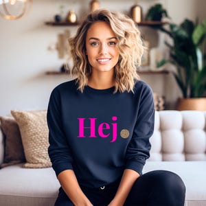 Könnte beinhalten: Ein marineblaues Sweatshirt mit dem Wort "Hej" in leuchtend pinkfarbenen Buchstaben. Rechts vom Wort befindet sich ein kleiner goldener Kreis mit Leopardenmuster. Das Sweatshirt hat einen Rundhalsausschnitt.