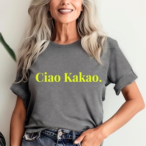 Pode incluir: Uma camiseta cinza com o texto "Ciao Kakao" em amarelo brilhante. A camiseta tem mangas enroladas e é feita de um material macio. A camiseta é combinada com jeans azuis.