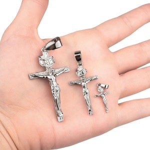 May include: Silver-colored crucifix pendants in three sizes. The pendants depict Jesus on the cross, with the inscription "INRI" above. The pendants are displayed on a hand, showcasing their size and detail.