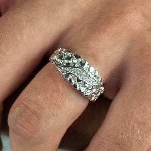 May include: A silver ring with a textured, nugget-like design. A central band of small, clear stones adds a sparkling accent. The ring is worn on a finger, showcasing its intricate details and craftsmanship.