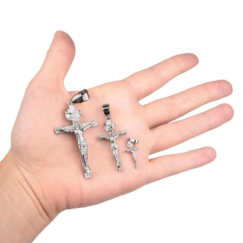 May include: Silver-colored crucifix pendants in three sizes. The pendants depict Jesus on the cross, with the inscription "INRI" above. The pendants are displayed on a hand, showcasing their size and detail.