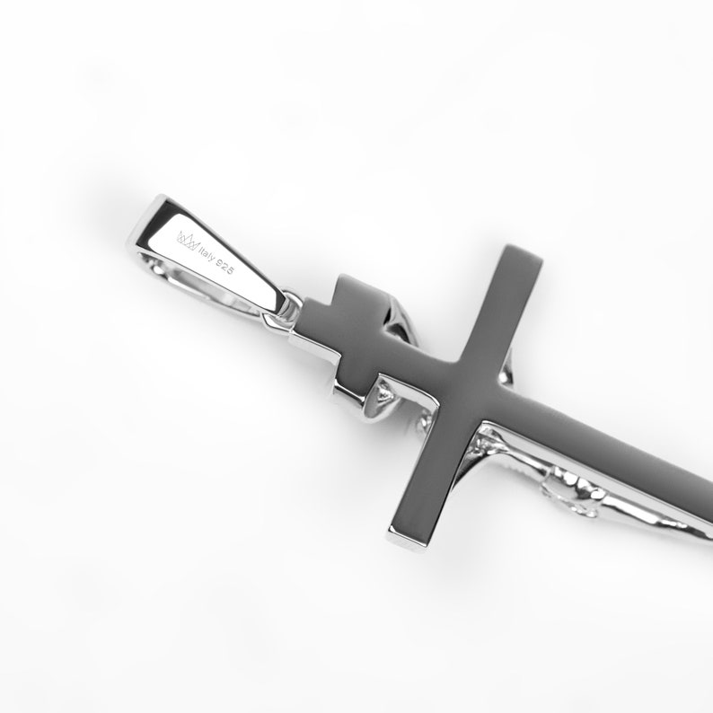 May include: A silver-colored pendant in the shape of a cross. The cross has a smooth, polished surface and a small loop at the top for a chain. The pendant is set against a white background.