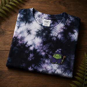 Può includere: Una t-shirt a maniche corte con motivo tie-dye nero, bianco e viola. Una toppa ricamata in basso a destra raffigura una rana verde con cappello e tunica da mago viola, che tiene in mano un bastone di legno. L'etichetta interna riporta Stiuka.
