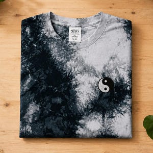 Könnte beinhalten: Ein kurzärmeliges T-Shirt mit Rundhalsausschnitt und einem schwarz-weißen Batikmuster. Das Shirt hat ein kleines gesticktes Yin-Yang-Symbol auf der Brust und ein weißes Innenetikett am Kragen mit dem in Schwarz gedruckten Text Shaka Wear TIE DYE.