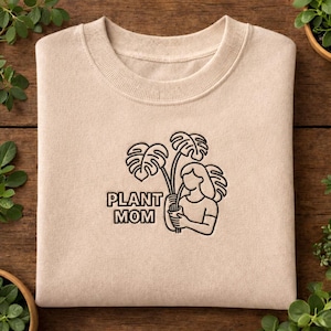 May include: A beige sweatshirt with a crew neck featuring a black line drawing of a person holding a plant with large leaves. The text "PLANT MOM" is below the illustration.