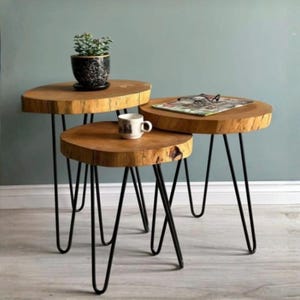 May include: Set of three wooden side tables with live edge tops and black hairpin legs. The tables are arranged in a nested configuration, with a small potted plant, a mug, and a magazine with glasses on top.
