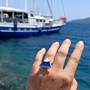 May include: A blue and white ceramic triangle ring with gold accents. The ring is on a hand, with a sailboat in the background.