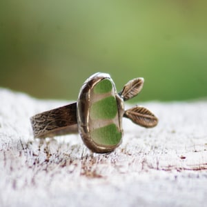 Könnte beinhalten: Ein silberner Ring mit einem grün-weißen Glasstein und zwei kleinen silbernen Blättern an der Seite.