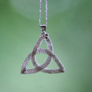 Puede incluir: Collar con colgante de nudo celta plateado. El colgante, un dise&ntilde;o triangular con bucles entrelazados, cuelga de una fina cadena. Un peque&ntilde;o detalle floral adorna la parte superior del colgante. El fondo es de un verde suave.