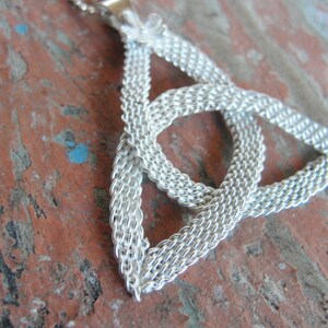 Puede incluir: Un collar plateado con un colgante de nudo celta. El colgante es un dise&ntilde;o tejido en forma triangular, con un peque&ntilde;o acento floral en la parte superior. La cadena es delicada y el fondo es una superficie r&uacute;stica roja y marr&oacute;n.