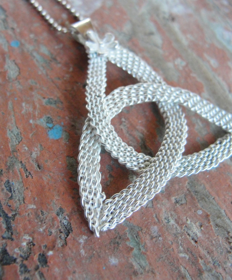Puede incluir: Un collar plateado con un colgante de nudo celta. El colgante es un dise&ntilde;o tejido en forma triangular, con un peque&ntilde;o acento floral en la parte superior. La cadena es delicada y el fondo es una superficie r&uacute;stica roja y marr&oacute;n.
