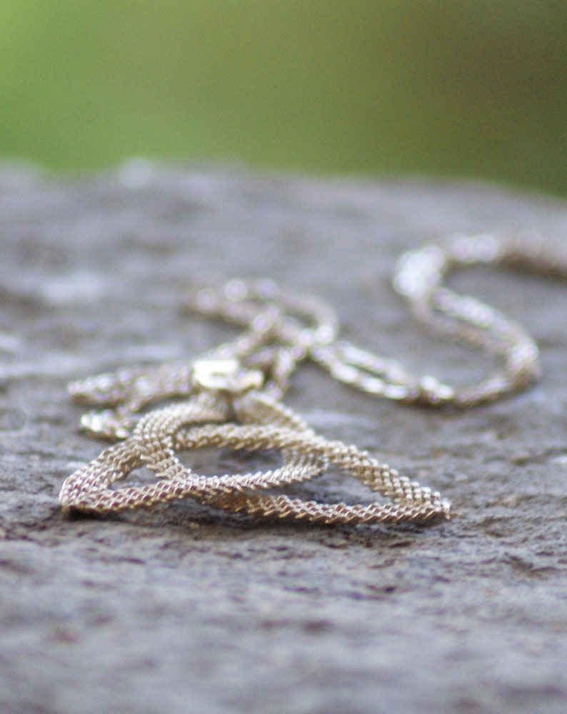 Puede incluir: Un delicado collar plateado con un dise&ntilde;o de cadena tejida. El collar est&aacute; sobre una superficie gris y texturizada, con un fondo verde borroso. La cadena tiene un dise&ntilde;o intrincado, reflejando la luz.