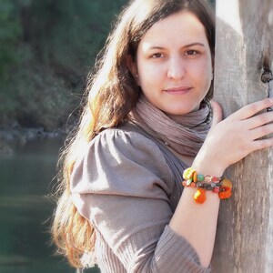 May include: A woman with long brown hair wearing a brown sweater and a scarf. She is wearing a colorful beaded bracelet on her wrist.