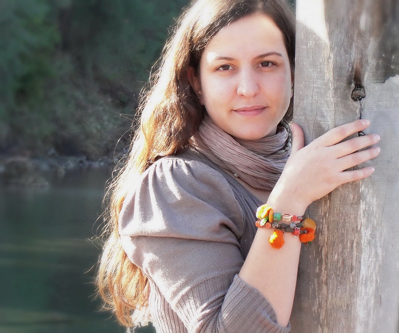 May include: A woman with long brown hair wearing a brown sweater and a scarf. She is wearing a colorful beaded bracelet on her wrist.
