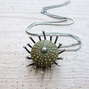 Puede incluir: Un collar de cadena de plata con un colgante de erizo de mar verde y negro. El colgante tiene una superficie texturizada y detalles plateados.