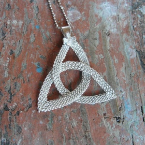 Puede incluir: Collar con colgante de nudo celta plateado. El colgante presenta un dise&ntilde;o triangular entrelazado y detallado, suspendido de una fina cadena. El collar se muestra sobre una superficie de madera envejecida de color marr&oacute;n rojizo.