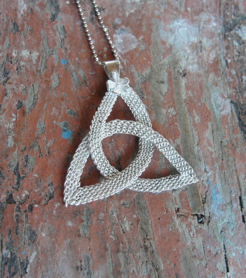 Puede incluir: Collar con colgante de nudo celta plateado. El colgante presenta un dise&ntilde;o triangular entrelazado y detallado, suspendido de una fina cadena. El collar se muestra sobre una superficie de madera envejecida de color marr&oacute;n rojizo.