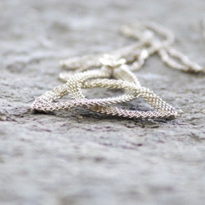 Puede incluir: Un collar plateado con una cadena delicada y un peque&ntilde;o colgante de flor. El collar est&aacute; sobre una superficie gris texturizada, con la cadena formando una forma triangular. El primer plano resalta el intrincado dise&ntilde;o tejido de la cadena.