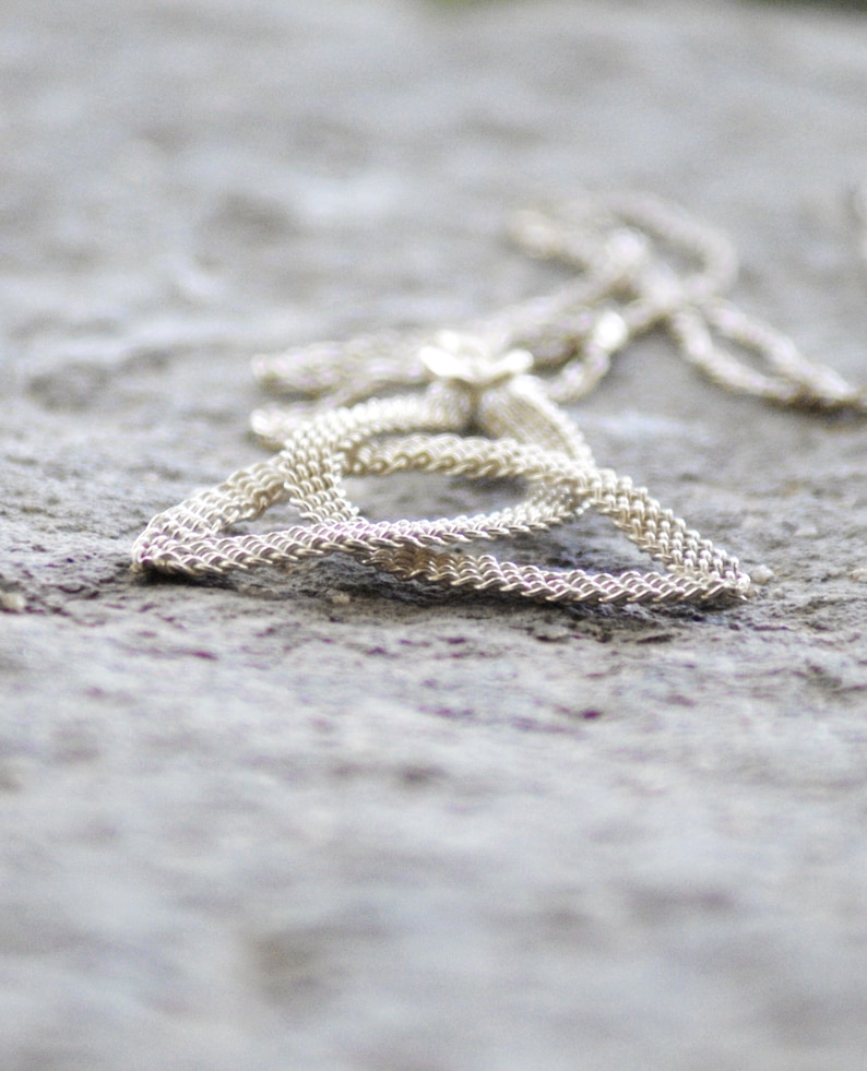 Puede incluir: Un collar plateado con una cadena delicada y un peque&ntilde;o colgante de flor. El collar est&aacute; sobre una superficie gris texturizada, con la cadena formando una forma triangular. El primer plano resalta el intrincado dise&ntilde;o tejido de la cadena.