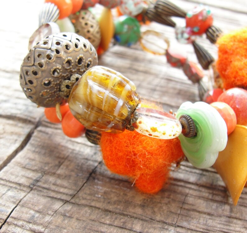 May include: A colorful bracelet with beads of various materials, including glass, metal, and fabric. The bracelet features a mix of orange, green, brown, and white beads.