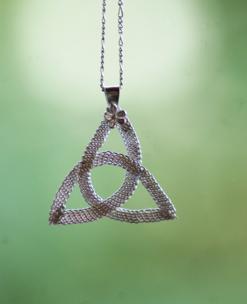 Puede incluir: Un collar con colgante plateado con un dise&ntilde;o de nudo celta. El colgante tiene forma triangular tejida con un peque&ntilde;o adorno floral en la parte superior, suspendido de una delicada cadena. El fondo es verde suave.