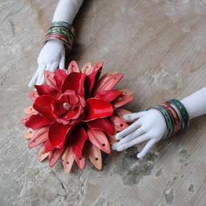 May include: A red and pink metal flower with a white center. The flower is surrounded by two white hands wearing colorful bracelets.