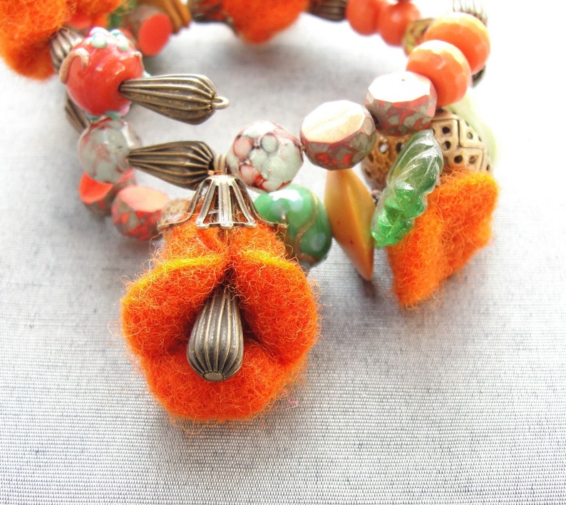 May include: A close-up of a colorful bracelet made with orange, green, and brown beads. The bracelet has a large orange felt flower charm attached to it.