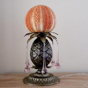 Puede incluir: Lámpara decorativa única con base en forma de piña y parte superior de erizo de mar. La lámpara presenta una base y detalles plateados, con colgantes de cristal rosa en forma de lágrima. Una pieza decorativa.
