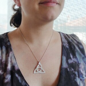 Puede incluir: Un collar plateado con un colgante de nudo celta. El colgante tiene forma triangular con un dise&ntilde;o entrelazado. El collar se lleva sobre una parte superior oscura estampada.