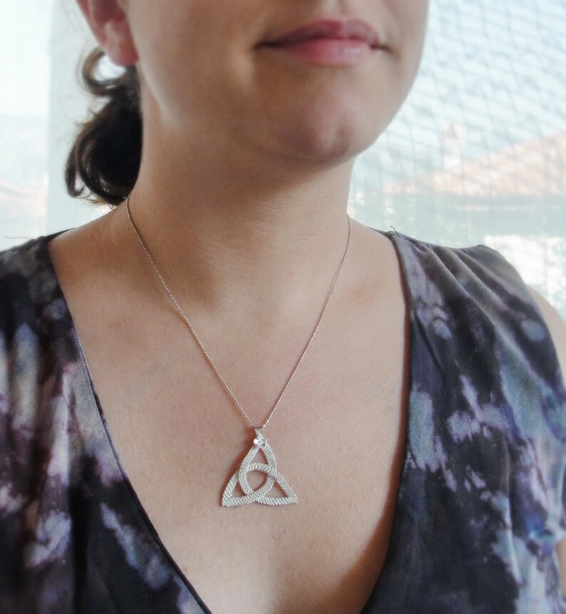 Puede incluir: Un collar plateado con un colgante de nudo celta. El colgante tiene forma triangular con un dise&ntilde;o entrelazado. El collar se lleva sobre una parte superior oscura estampada.