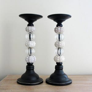 May include: Pair of black candle holders with a decorative design. The candle holders have a round base, a stem with clear glass beads and white sea urchin accents, and a black top. The candle holders are displayed on a wooden surface.