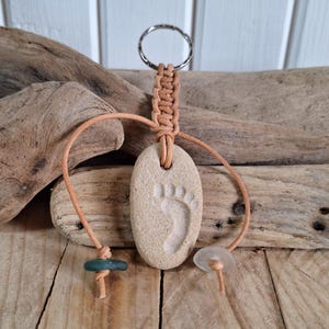 Op de afbeelding: Een beige stenen sleutelhanger met een uitgesneden voetafdruk. De sleutelhanger heeft een gevlochten bruin koord dat aan een metalen ring is bevestigd. Twee kleine kralen, een groene en een transparante, zijn aan het koord bevestigd. De achtergrond bestaat uit drijfhout en houten planken.