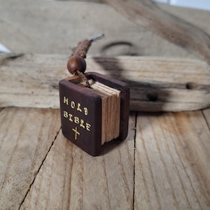 Könnte beinhalten: Ein kleiner Holzbuch-Anhänger mit braunem Einband und goldenen Lettern, auf dem "HOLY BIBLE" mit einem goldenen Kreuz unter dem Text steht.