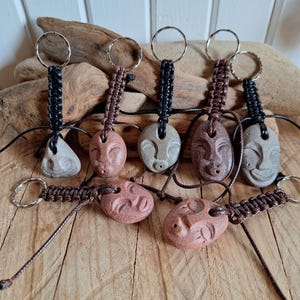 Op de afbeelding: Een verzameling van zeven sleutelhangers met gladde, uitgesneden stenen met mensachtige gezichtsuitdrukkingen. Elke steen is bevestigd aan een zwart of bruin gevlochten koord, dat verbonden is met een ronde metalen sleutelring in diverse kleuren.