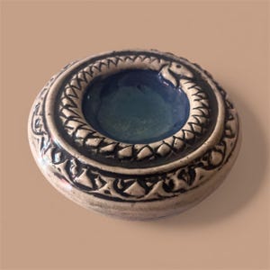 May include: A ceramic ashtray with a circular design. The ashtray features a blue interior and a raised border with a snake motif. The exterior is a cream color with black decorative patterns. The ashtray is round and has a small footprint.