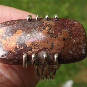 May include: A ring with a large, irregularly shaped, brown and white stone with copper flecks. The stone is set in a silver band with five prongs.