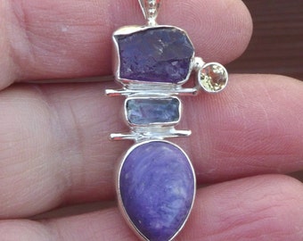 May include: A silver pendant with a layered design featuring four different gemstones. The top gemstone is a purple amethyst, followed by a blue aquamarine, a smaller purple amethyst, and a yellow citrine at the top.