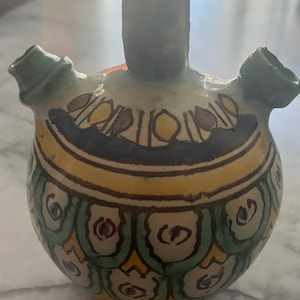 Peut inclure: Une cruche en céramique avec un motif géométrique vert, jaune et bleu. La cruche a un corps large et rond et un col étroit avec un bec de chaque côté.