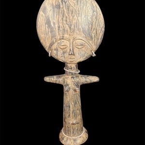 May include: A carved wooden figure with a large, round head and a stylized face. The figure has outstretched arms and a cylindrical body. It is painted in shades of brown and beige, with a textured surface. Small white beads adorn the neck and ears.