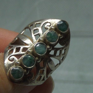 May include: A silver ring with a filigree design and five green gemstones. The ring is on a finger.